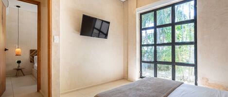 A bedroom with a bed, a TV mounted on the wall, and a large window with a view of palm trees.