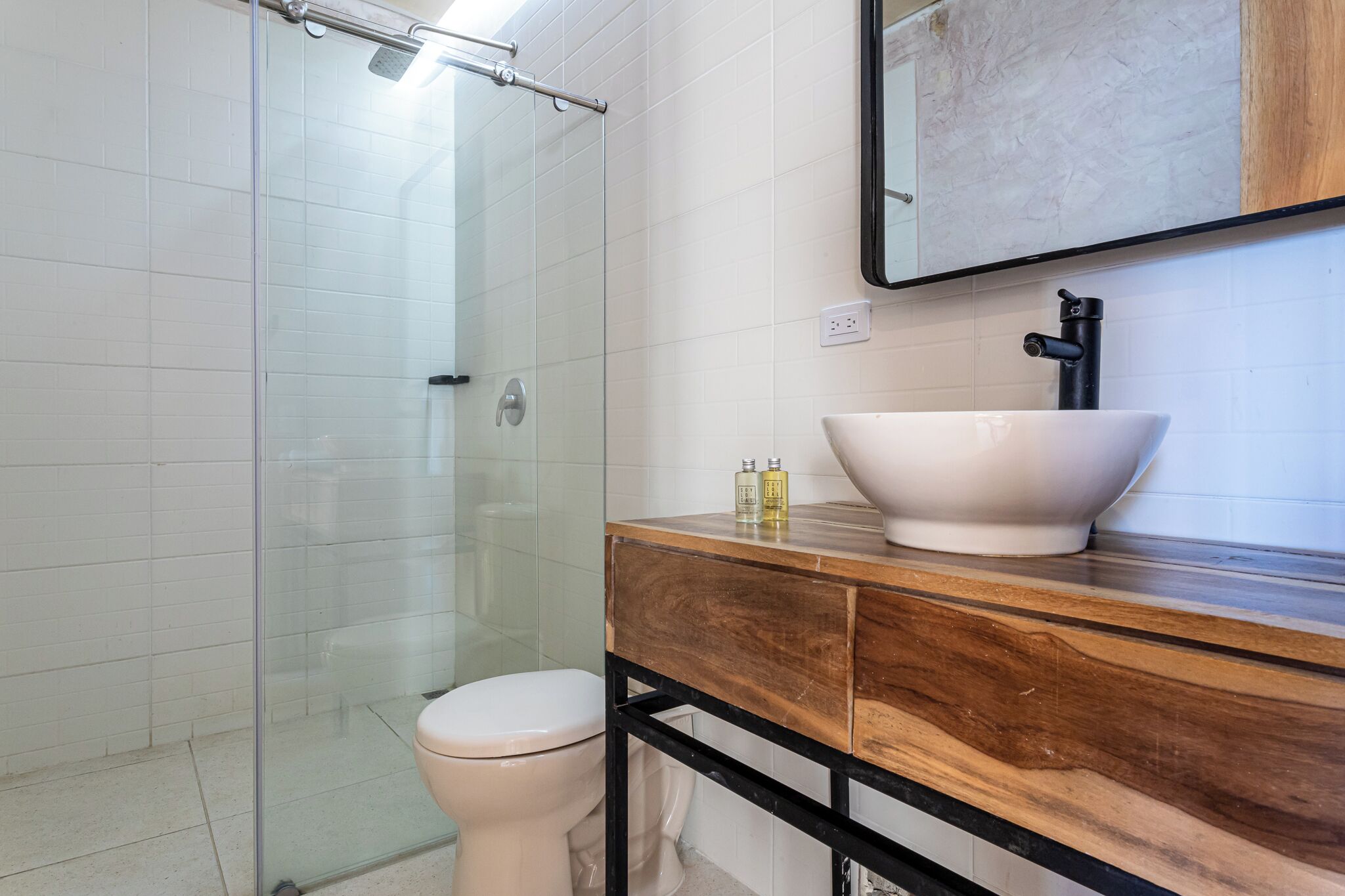 A bathroom with a glass shower enclosure, a white toilet, a wooden vanity with a white sink, and a mirror above the sink.