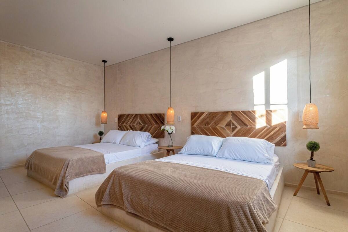 A hotel room with two beds, a wooden headboard, a small round table with a plant, and pendant lights.