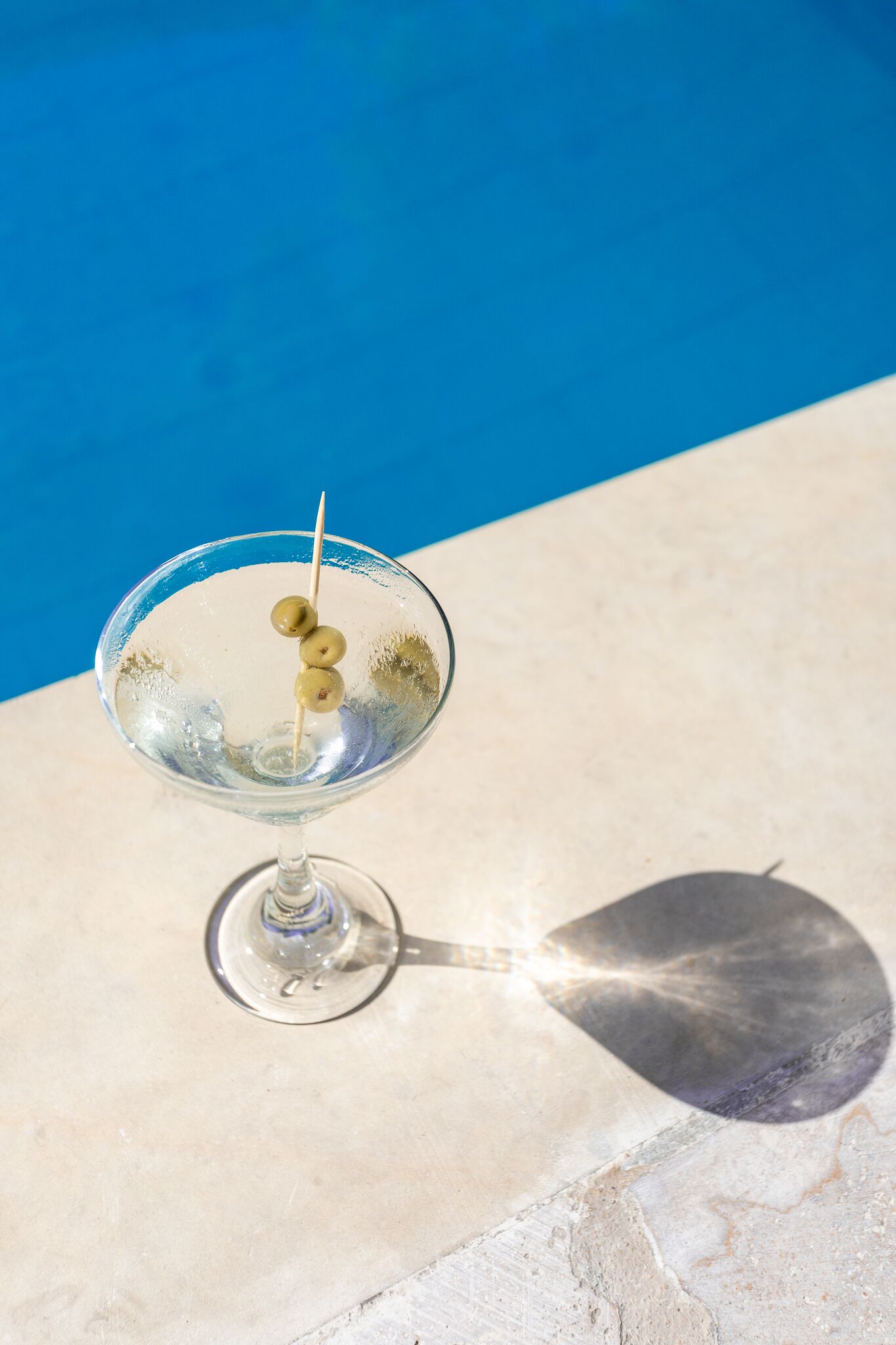 A clear glass martini glass with three olives on a reflective surface near a pool.