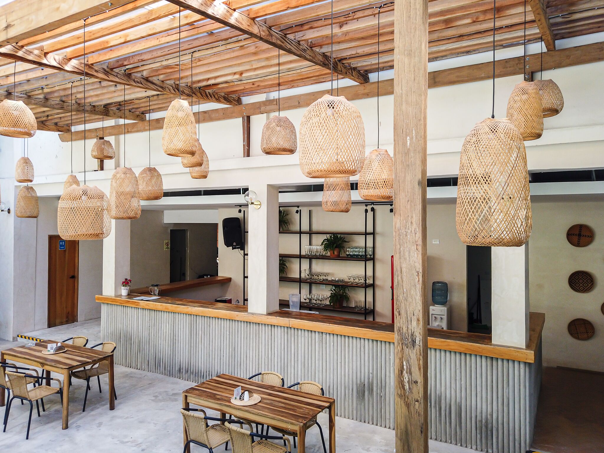 A restaurant with wooden tables and chairs, hanging woven light fixtures, and a counter with shelves of dishes and glasses.