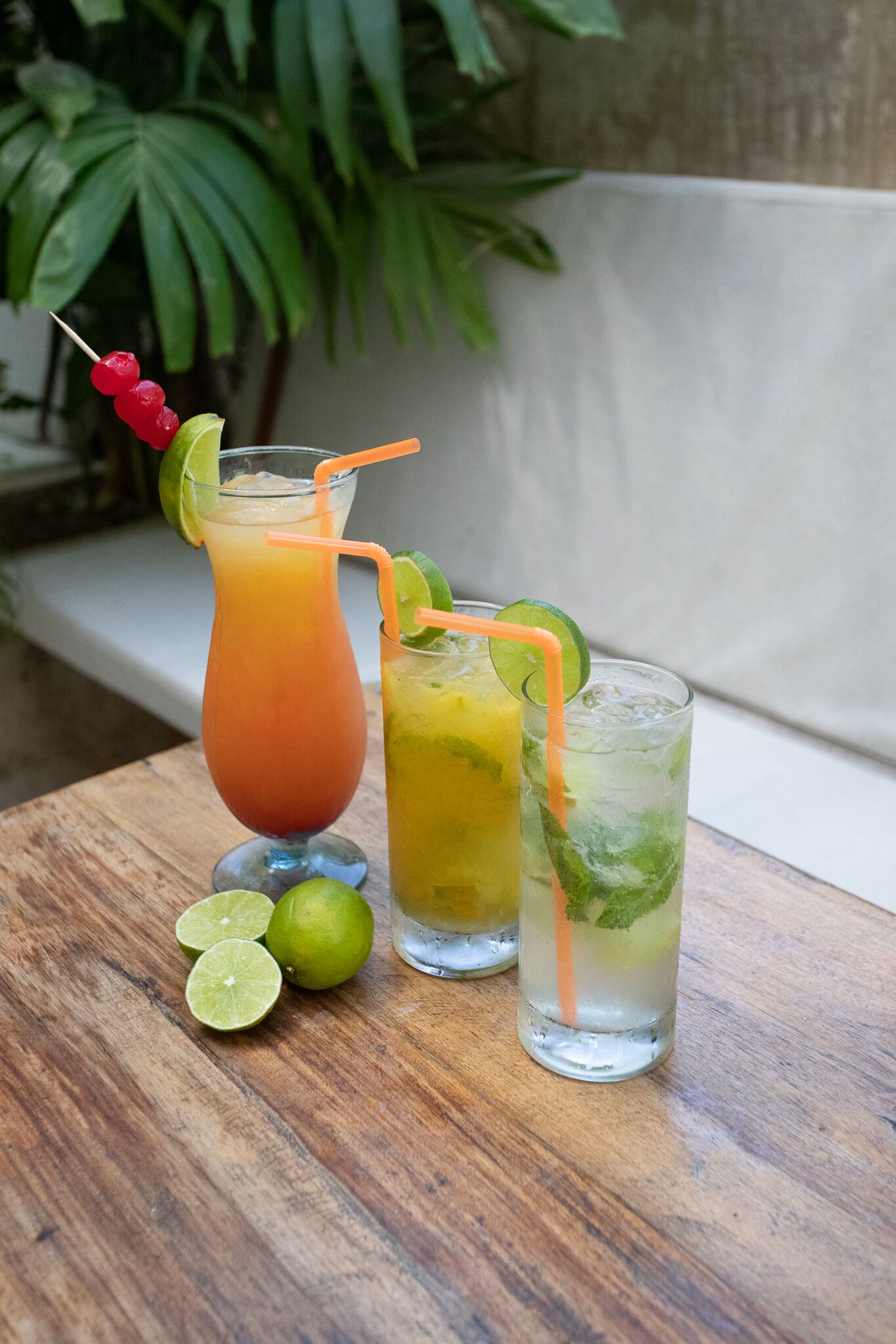 Two drinks on a wooden table with lime wedges and a plant in the background.