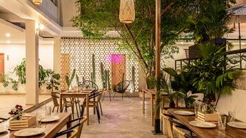 An indoor dining area with wooden tables and chairs, surrounded by potted plants and a decorative fence.