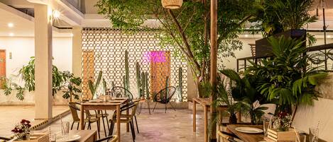 An indoor dining area with wooden tables and chairs, surrounded by potted plants and a decorative fence.