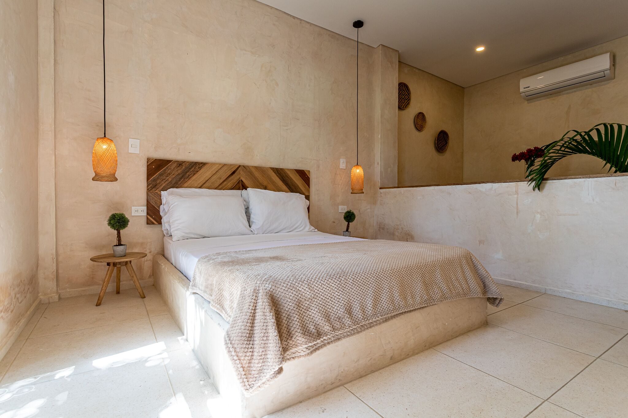 Un dormitorio con cama, dos mesitas de noche, una planta pequeña en maceta y un aire acondicionado instalado en la pared.