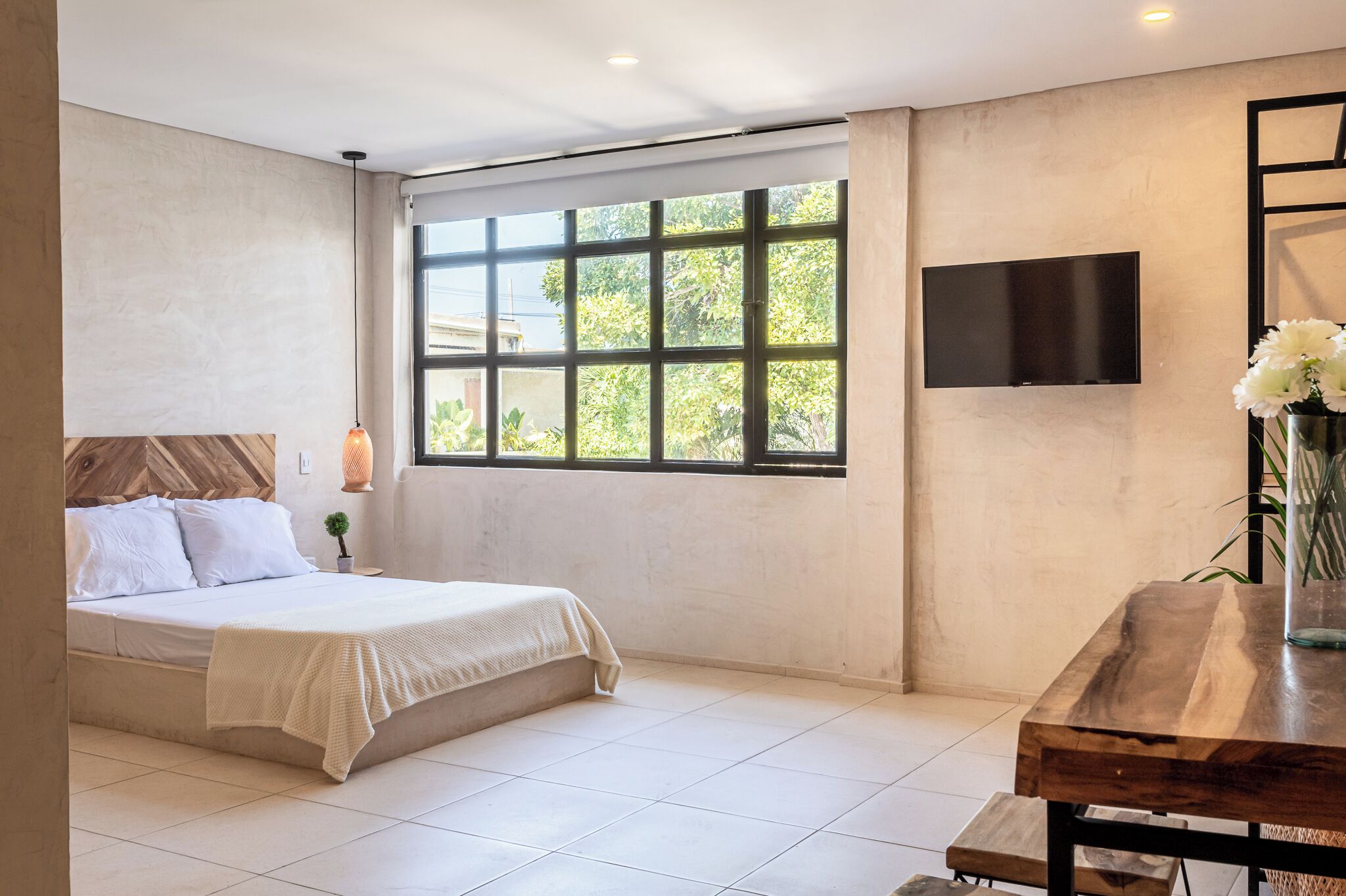 A bedroom with a bed, a TV, a window with a view of greenery, and a wooden table with a vase.