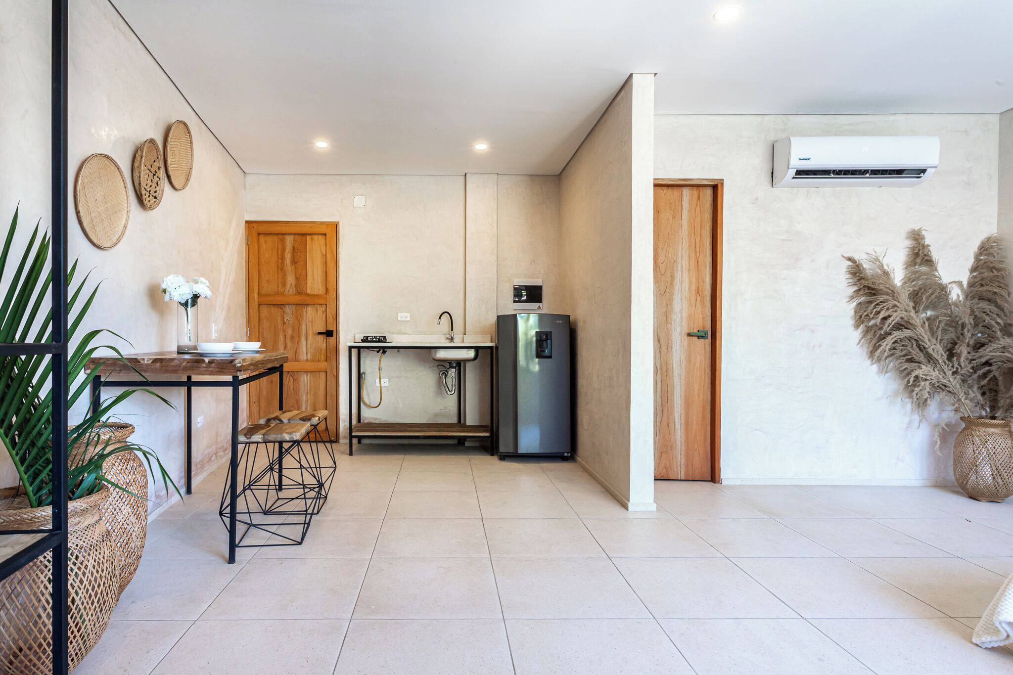 A modern kitchen with a wooden door, a built-in sink, and a stainless steel refrigerator. There is a dining table with chairs and a vase of flowers.