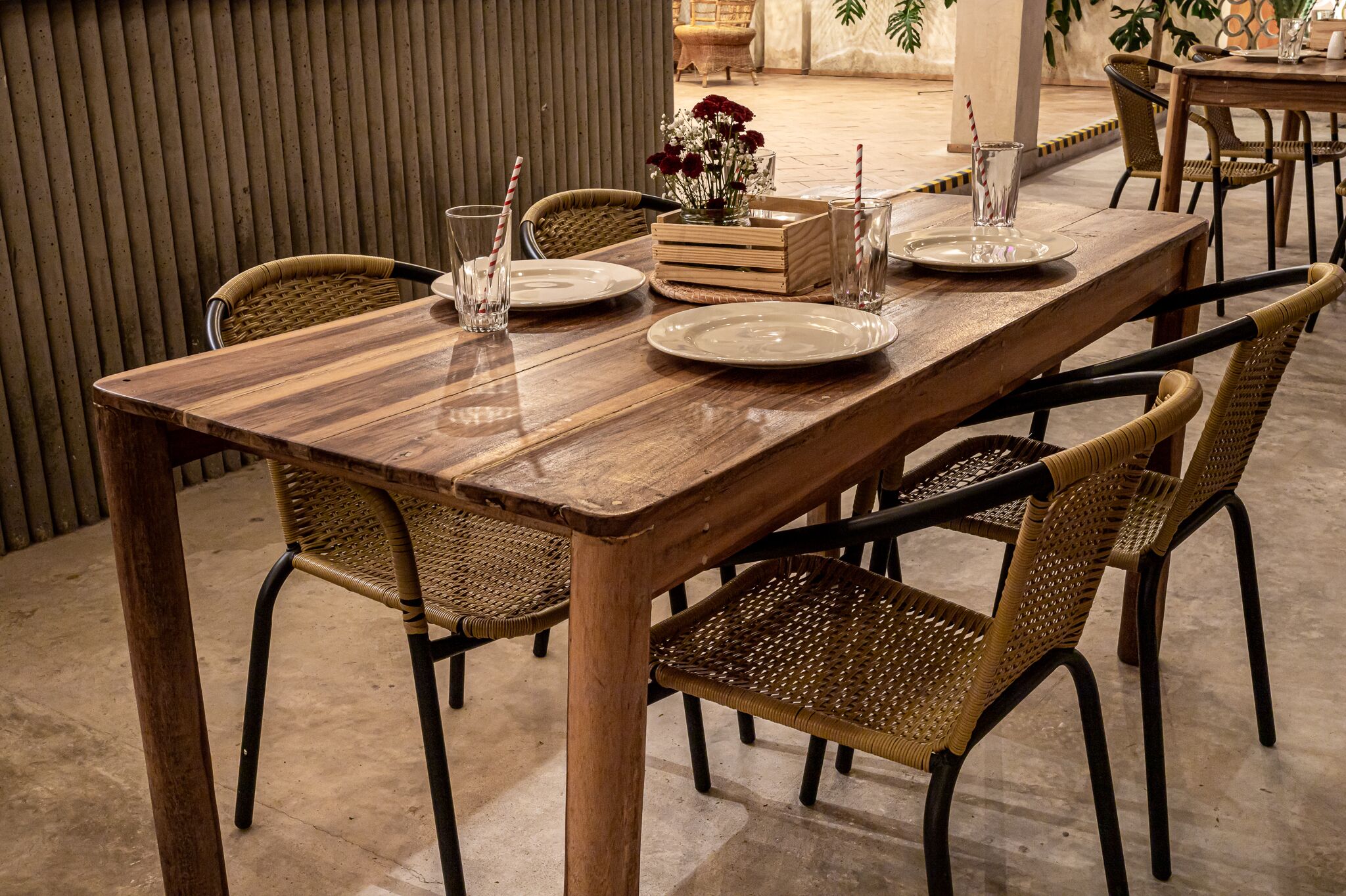 A wooden dining table with four wicker chairs, a vase with flowers, and two glasses with straws.