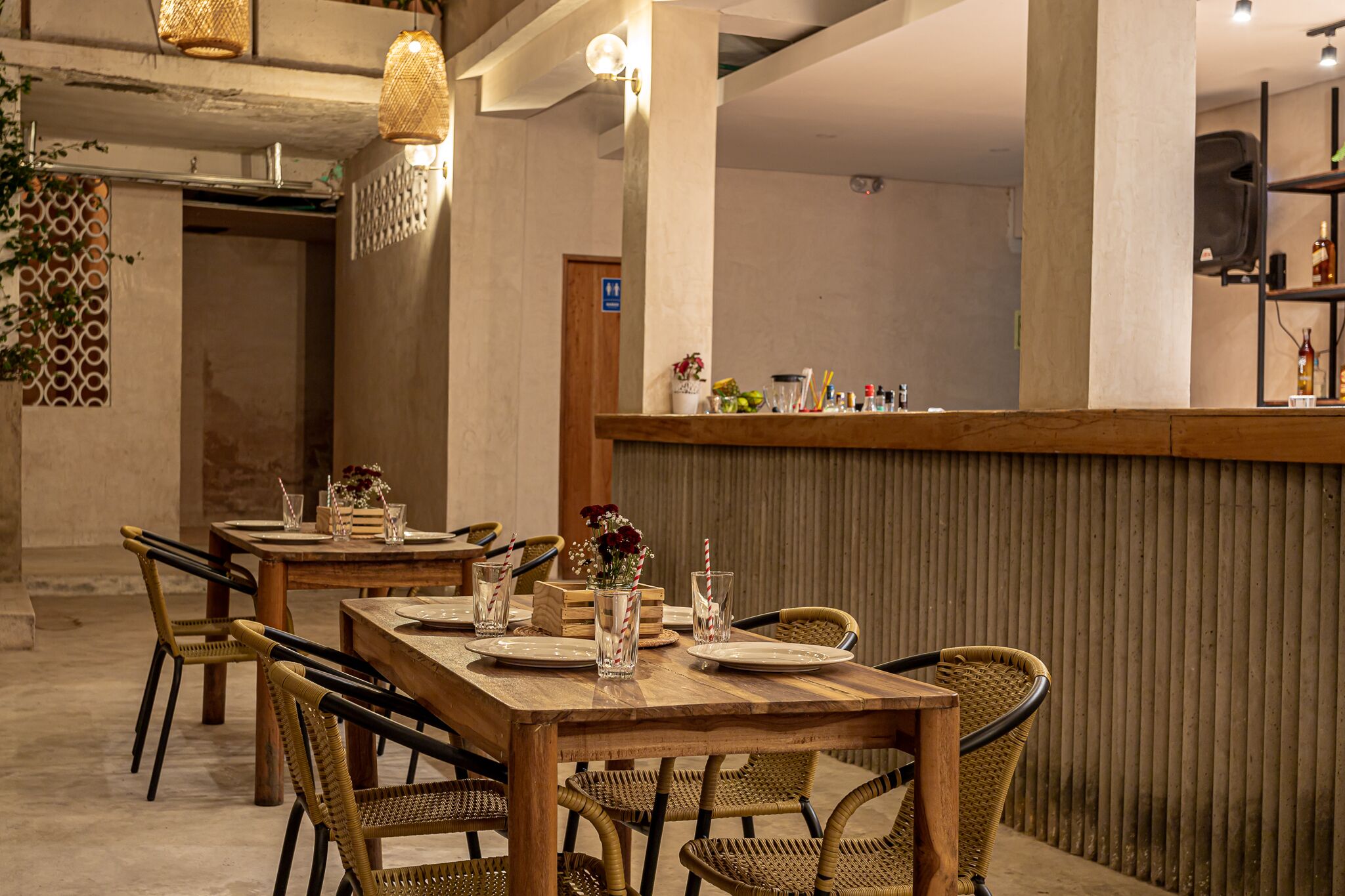 A restaurant with wooden tables and wicker chairs, a bar area with bottles and glasses, and a decorative wall with a plant.