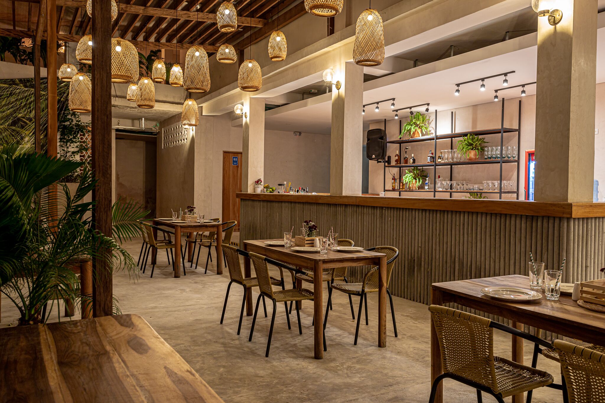 A restaurant interior with wooden tables and chairs, hanging wicker lamps, and a bar area with shelves of glassware and plants.