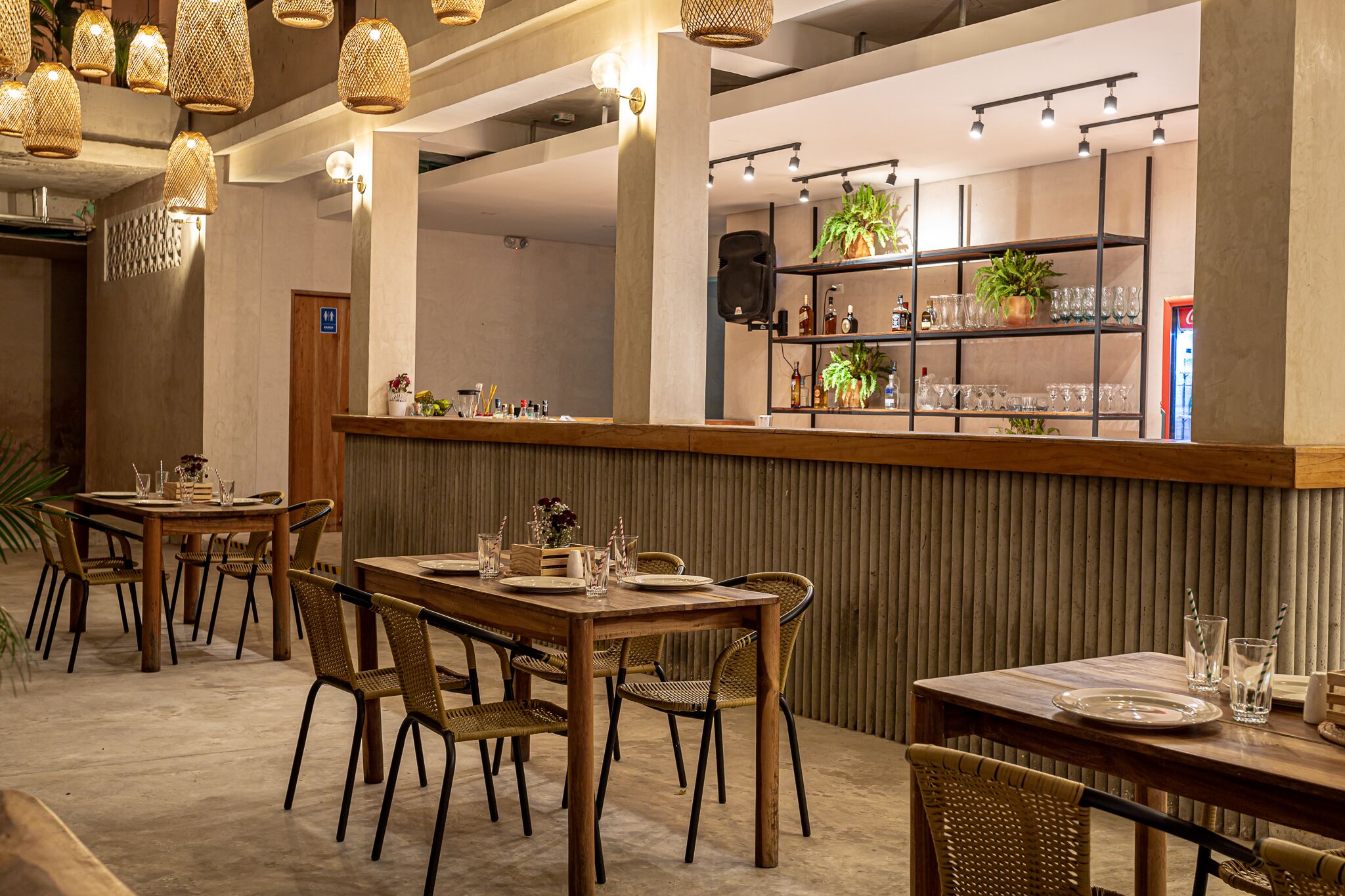 Ein Restaurantinneres mit Holztischen und -stühlen, einer Bar mit Regalen voller Gläser und Pflanzen, sowie hängenden Korbschwingenlampen.