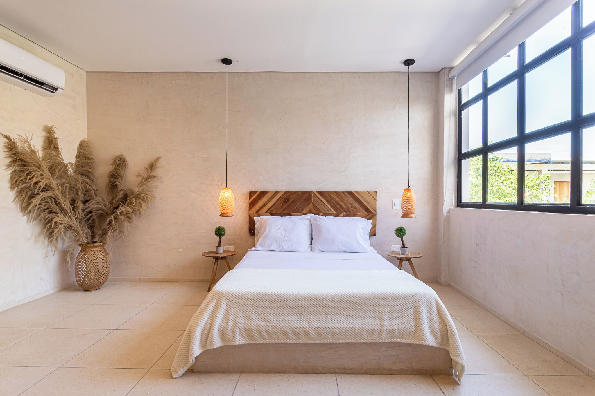A modern bedroom with a large bed, wooden headboard, hanging pendant lights, and a view through a large window.