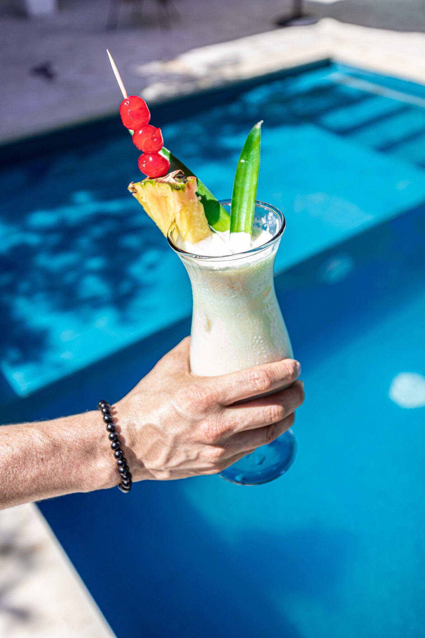 Una mano che regge un cocktail con uno spiedino di ciliegie e una fetta di ananas, vicino a una piscina.