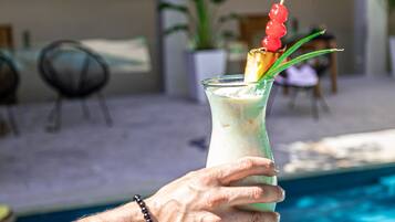 A hand holding a cocktail with a pineapple skewer, near a swimming pool with outdoor seating and plants.