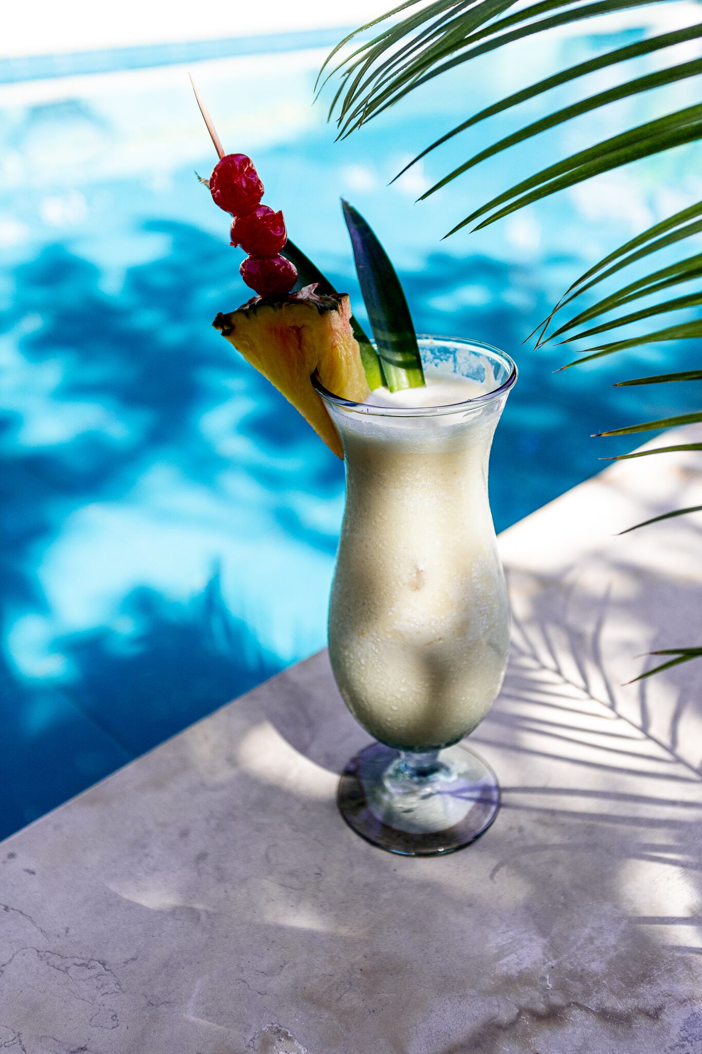 Un cocktail agrémenté d’une brochette d’ananas et d’une feuille décorative, posé sur une table au bord d’une piscine, avec un fond bleu clair.