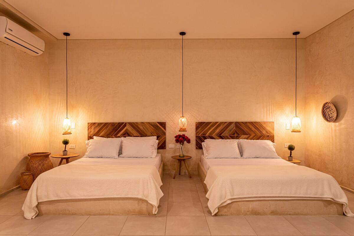 A hotel room with two beds, a wooden headboard, a small round table with a lamp, and a wall-mounted air conditioner.