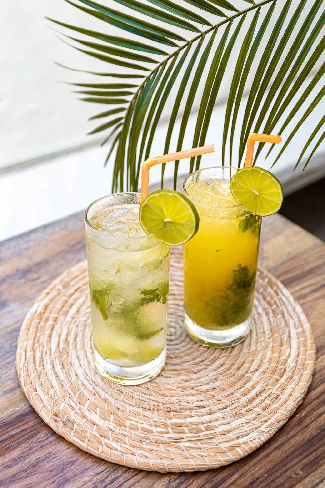 Two glasses of iced drinks with lime slices and straws on a woven mat.