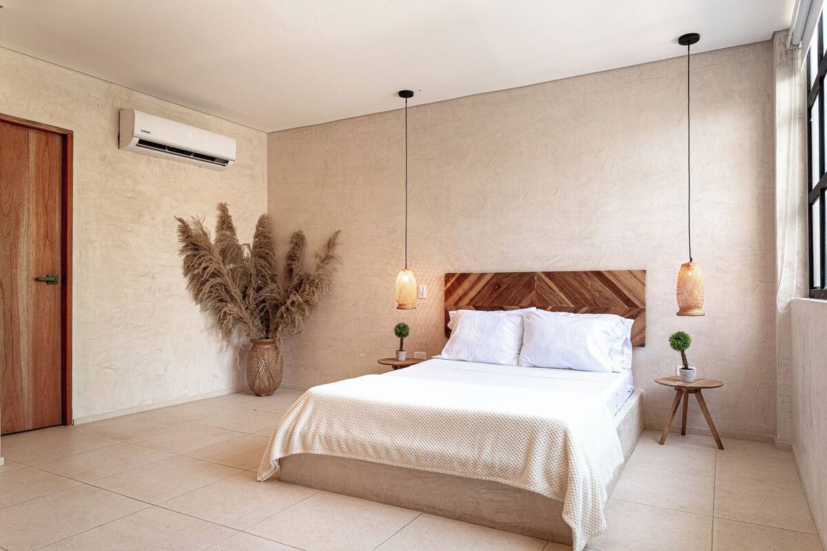 A bedroom with a bed, two bedside tables, a potted plant, and a hanging light fixture.