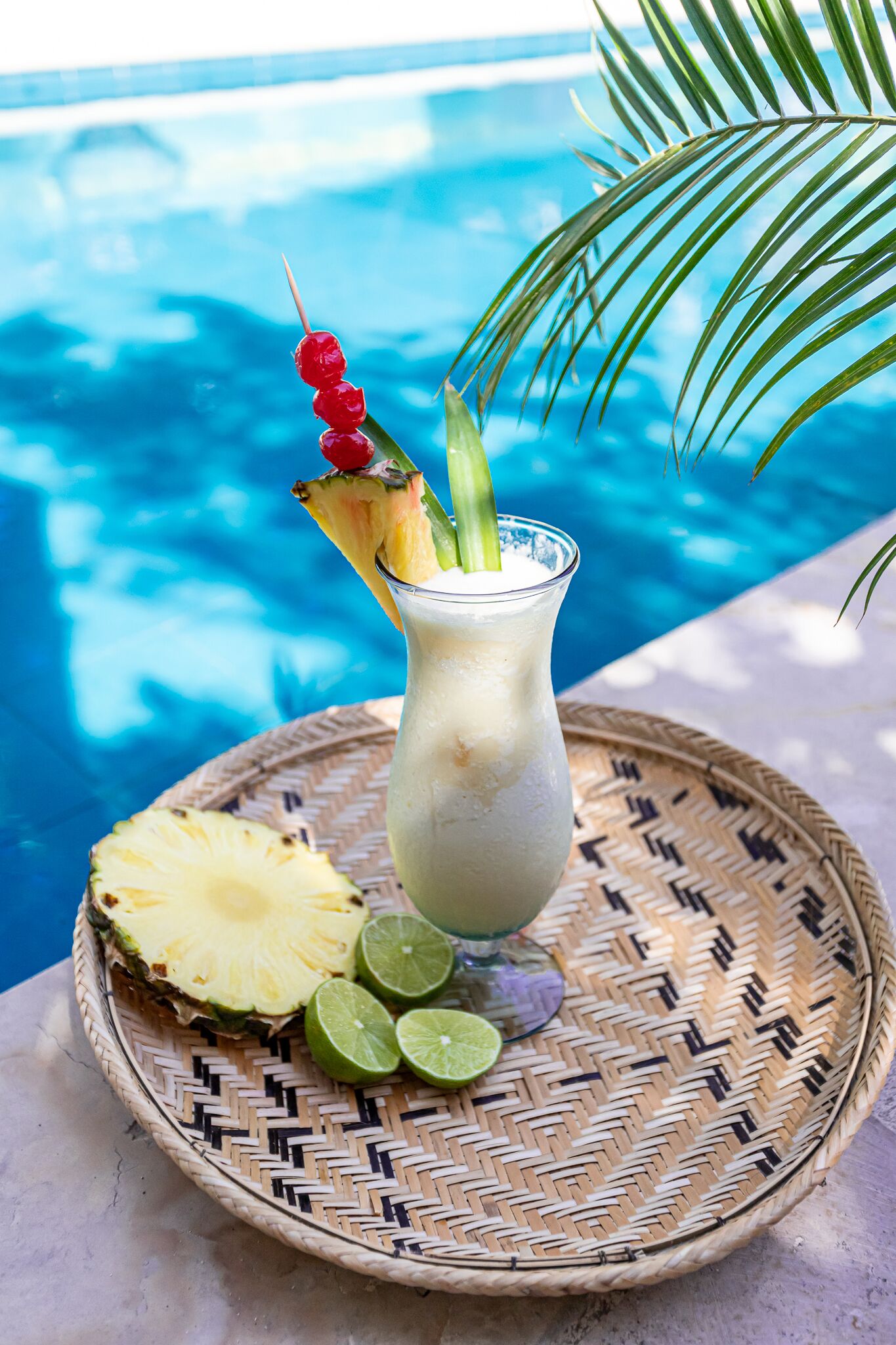 Una bevanda tropicale con una fetta di ananas e spicchi di lime su un tappetino di giunchi accanto a una piscina.