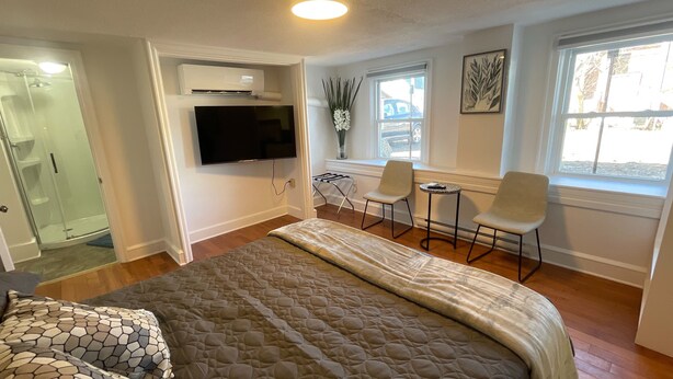 A bedroom with a large bed, a TV mounted on the wall, a window with a view, and a seating area with two chairs and a small table. - Studio-like Comfort in Wolfville’s Beating Heart (Wolfville)
