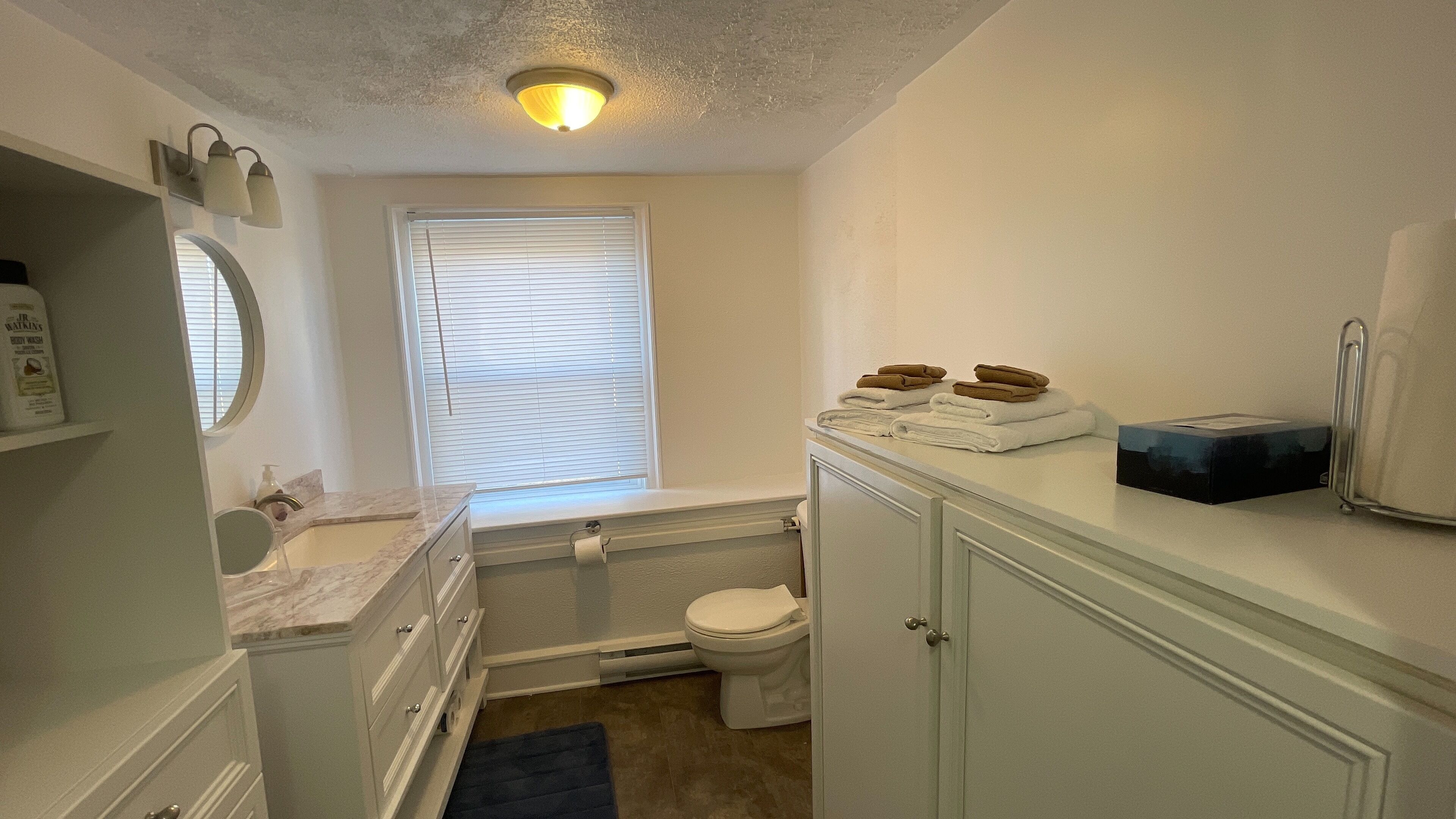 Baño pequeño con inodoro, lavamanos y espejo. Hay una ventana con persianas, un perchero con toallas y un dispensador de jabón en la encimera.