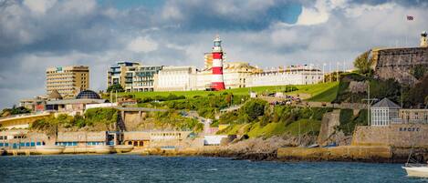 A coastal cityscape with a prominent lighthouse, a large white building with flags, and a rocky shoreline.
