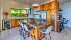 A modern kitchen with wooden cabinets, a central island with bar stools, stainless steel appliances, and a large window overlooking greenery. - Casa Privado - Hermosa Resort Villa #4 Costa Rica (Uvita)