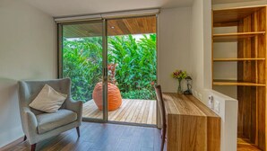 A modern room with a sliding glass door leading to a wooden deck, a grey armchair, a wooden desk, and a built-in shelving unit. - Casa Privado - Hermosa Resort Villa #4 Costa Rica (Uvita)