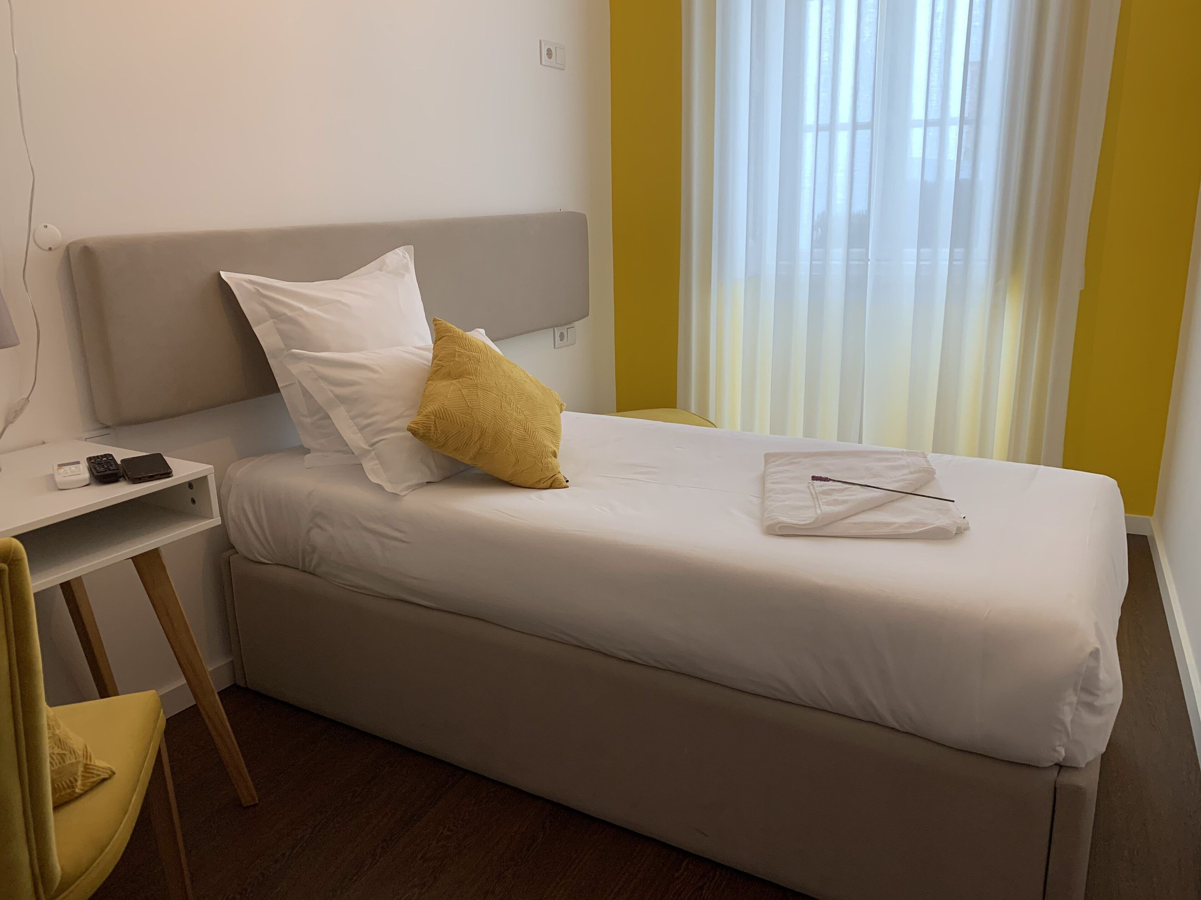 Ein ordentlich bezogenes Bett mit weißen Laken, einem gelben Kissen und einem gefalteten Handtuch. Ein kleiner Nachttisch mit Telefon und Geldbörse ist zu sehen. Der Raum hat ein schlichtes, sauberes Design mit Holzboden und einem Fenster mit Vorhängen.