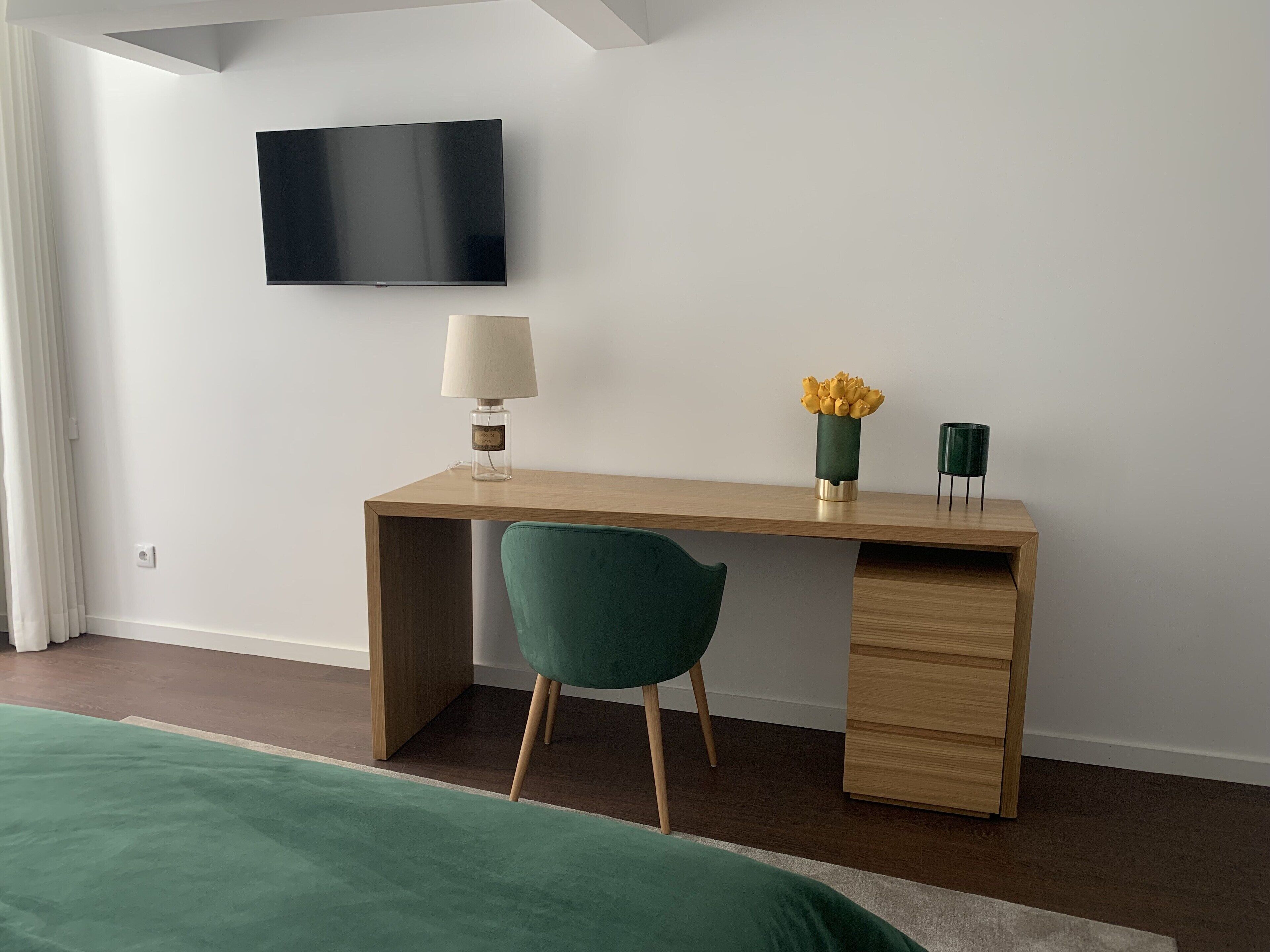 Un dormitorio moderno con un escritorio de madera, una silla verde, un televisor montado en la pared y una mesita de noche con una lámpara y flores.