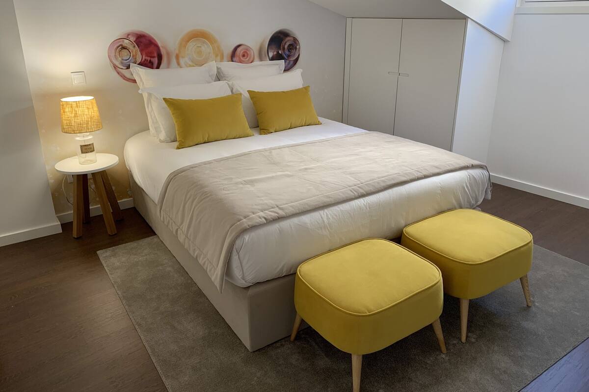 A bedroom with a bed, bedside table, lamp, and two yellow ottomans.