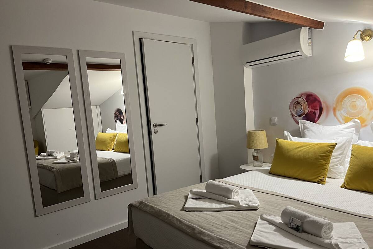 A hotel room with a bed, a mirror, a bedside table with a lamp, and an air conditioning unit.