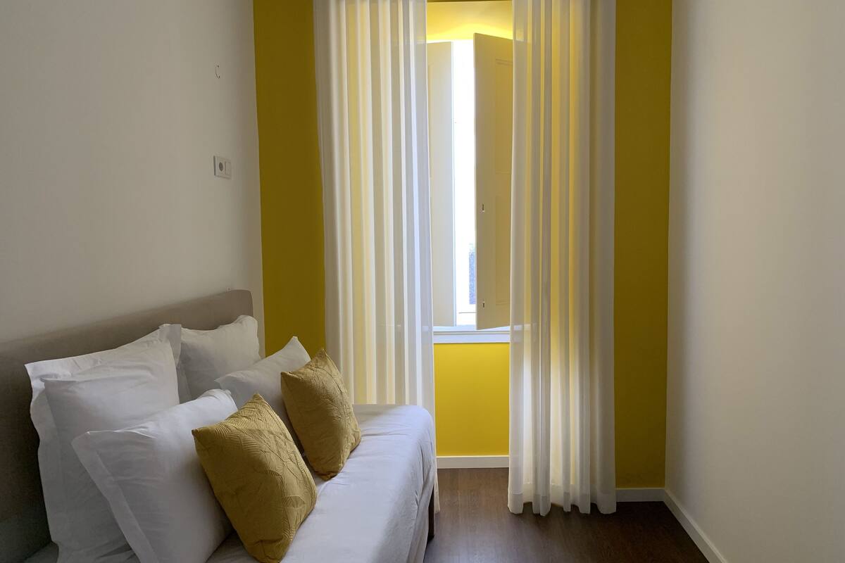 A room with a bed, white and mustard pillows, yellow walls, and a window with white curtains.