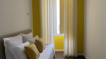 A room with a bed, white and mustard pillows, yellow walls, and a window with white curtains.