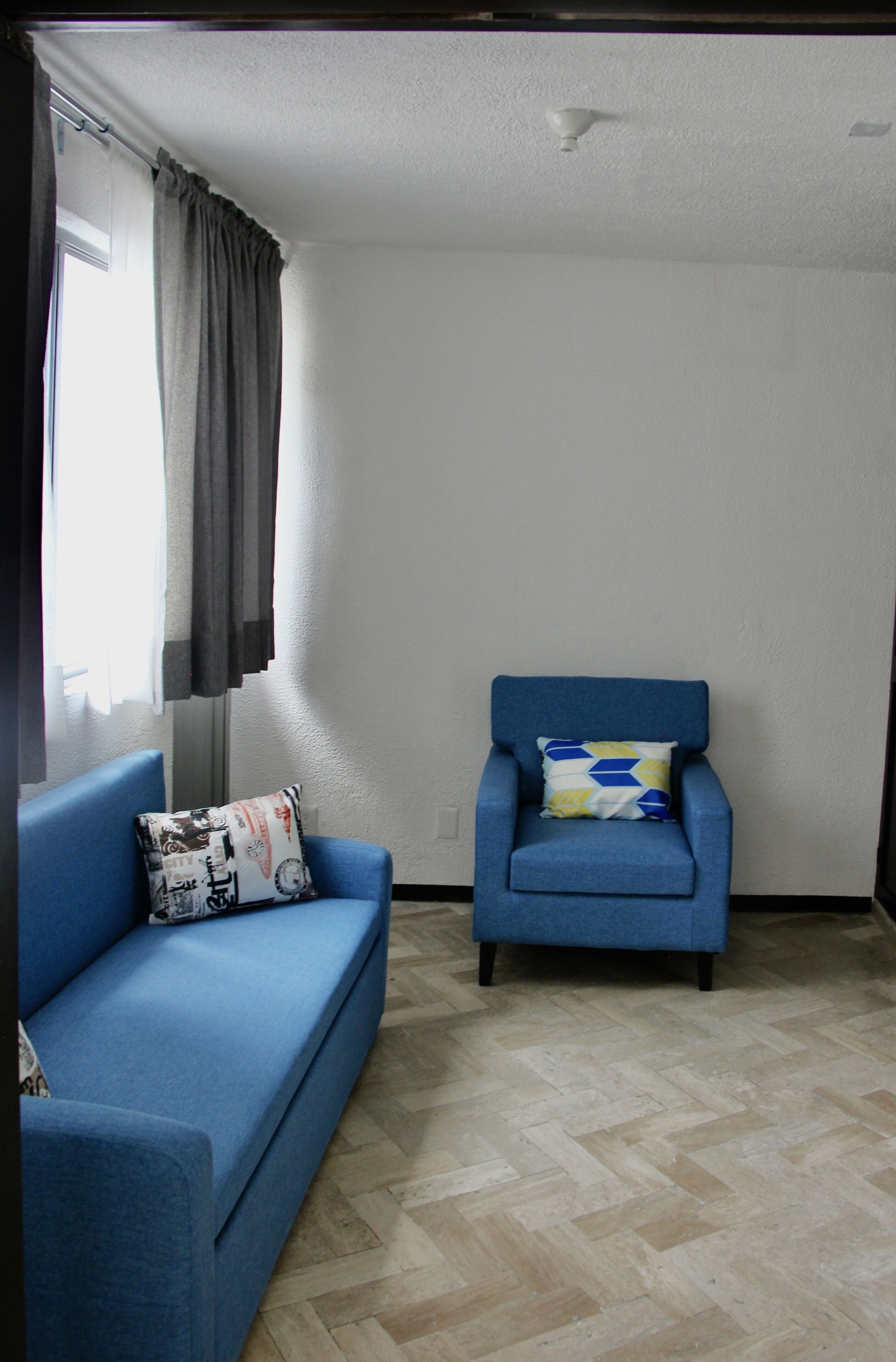 A room with a blue sofa and armchair, a window with curtains, and a white wall.