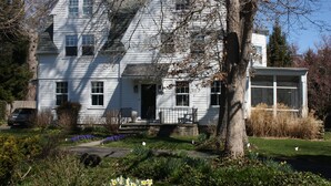 Une maison blanche à deux étages, dotée d’un porche à l’avant, d’un garage et d’un jardin où poussent diverses plantes et fleurs.