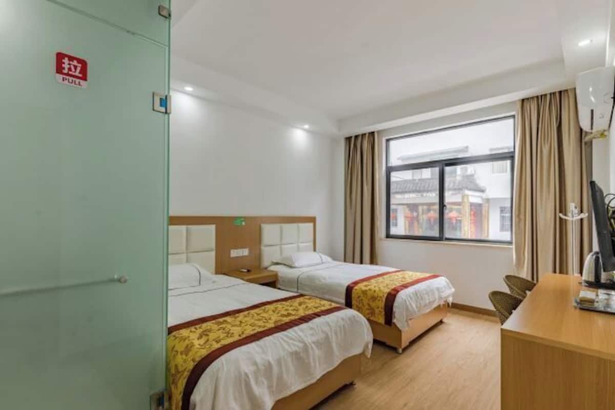 A hotel room with two beds, a desk, a chair, a window with curtains, and a visible door with a pull sign.