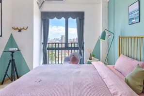 A bedroom with a bed, a window with curtains, a desk with a lamp, and a city view.