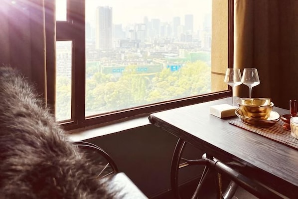 A hotel room with a city view, a dining table with a gold bowl and wine glasses, and a fur throw on the chair.