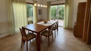Ein Esszimmer mit Holztisch und -stühlen, großen Fenstern mit Vorhängen und Blick ins Grüne.