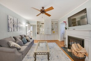 Ein Wohnzimmer mit grauem Sofa, einem Glastisch, einem Kamin und einem Deckenventilator.