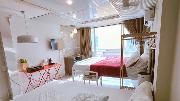 Ein Hotelzimmer mit einem großen Bett, einem Schreibtisch und einem Stuhl. Es gibt ein Fenster mit Vorhängen, einen Deckenventilator und einen an der Wand befestigten Fernseher.