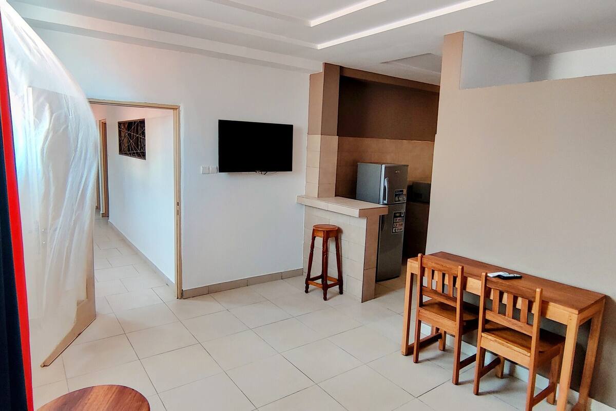 Un espacio habitable compacto con kitchenette, una zona de comedor con mesa y sillas de madera, un televisor montado en la pared y un refrigerador.