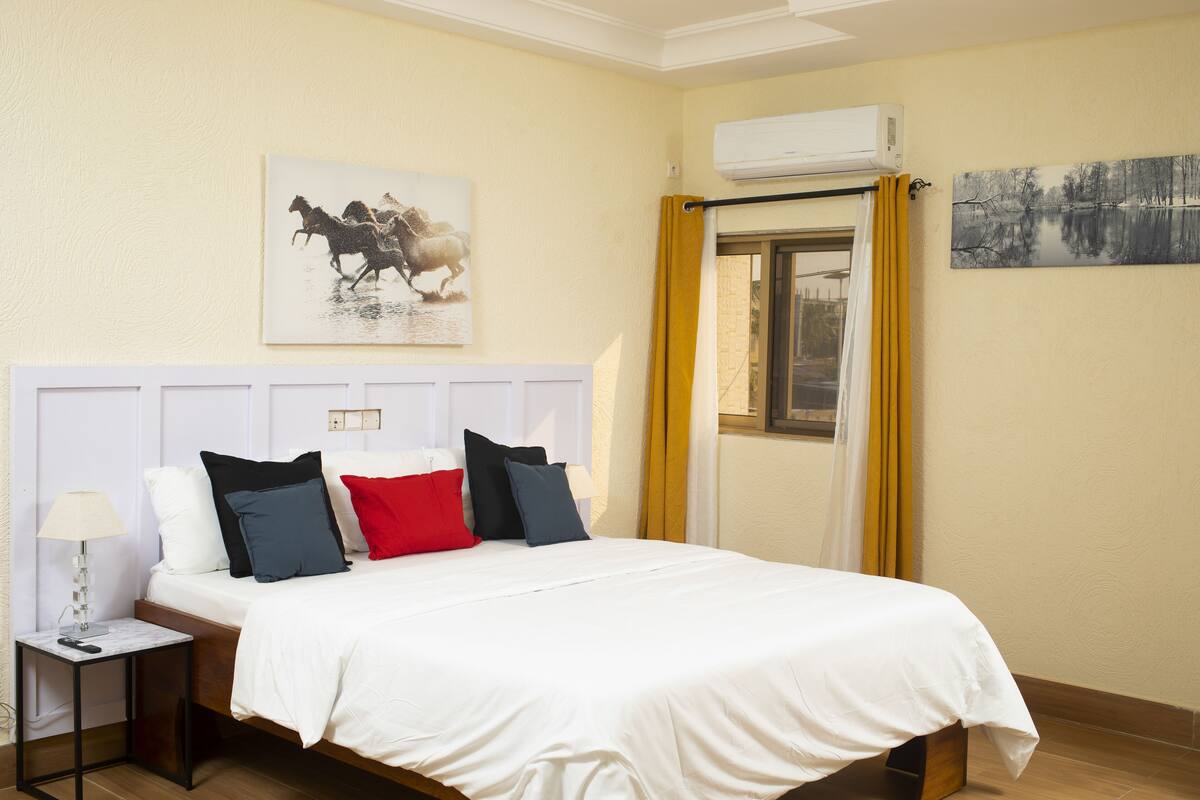 Habitación de hotel con cama, mesita de noche, lámpara, ventana con cortinas y dos cuadros colgados en la pared.