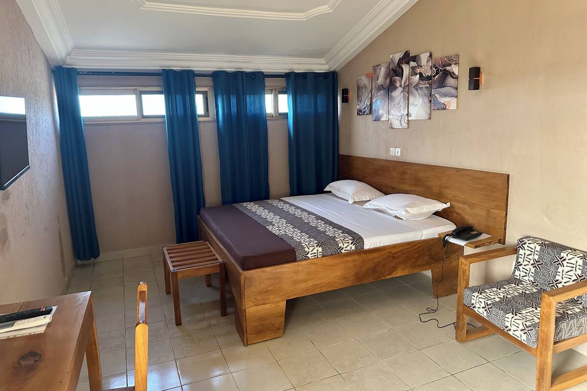Habitación de hotel con cama de madera, mesita de noche de madera, una silla con cojín estampado, una pequeña mesa de madera y un televisor montado en la pared.