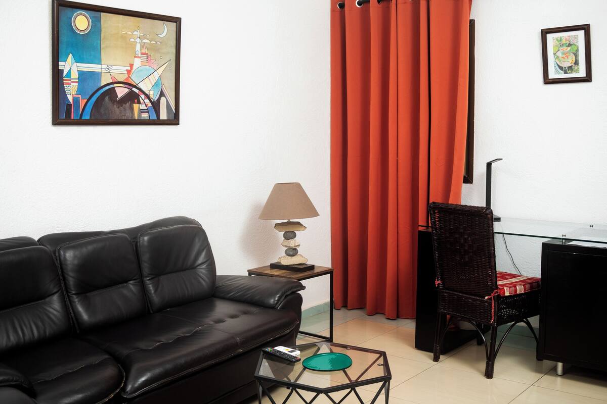 Una sala de estar con un sofá de cuero negro, una mesa de centro de vidrio, una lámpara, un sillón de mimbre, una cortina roja y un cuadro en la pared.