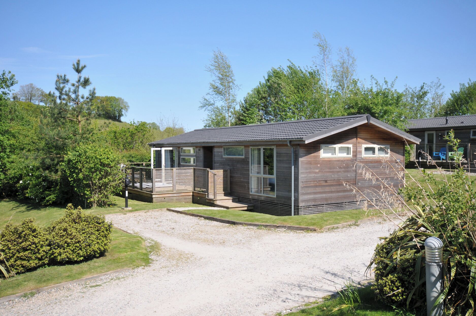 A small wooden cabin with a porch, a gravel driveway, and surrounding greenery.