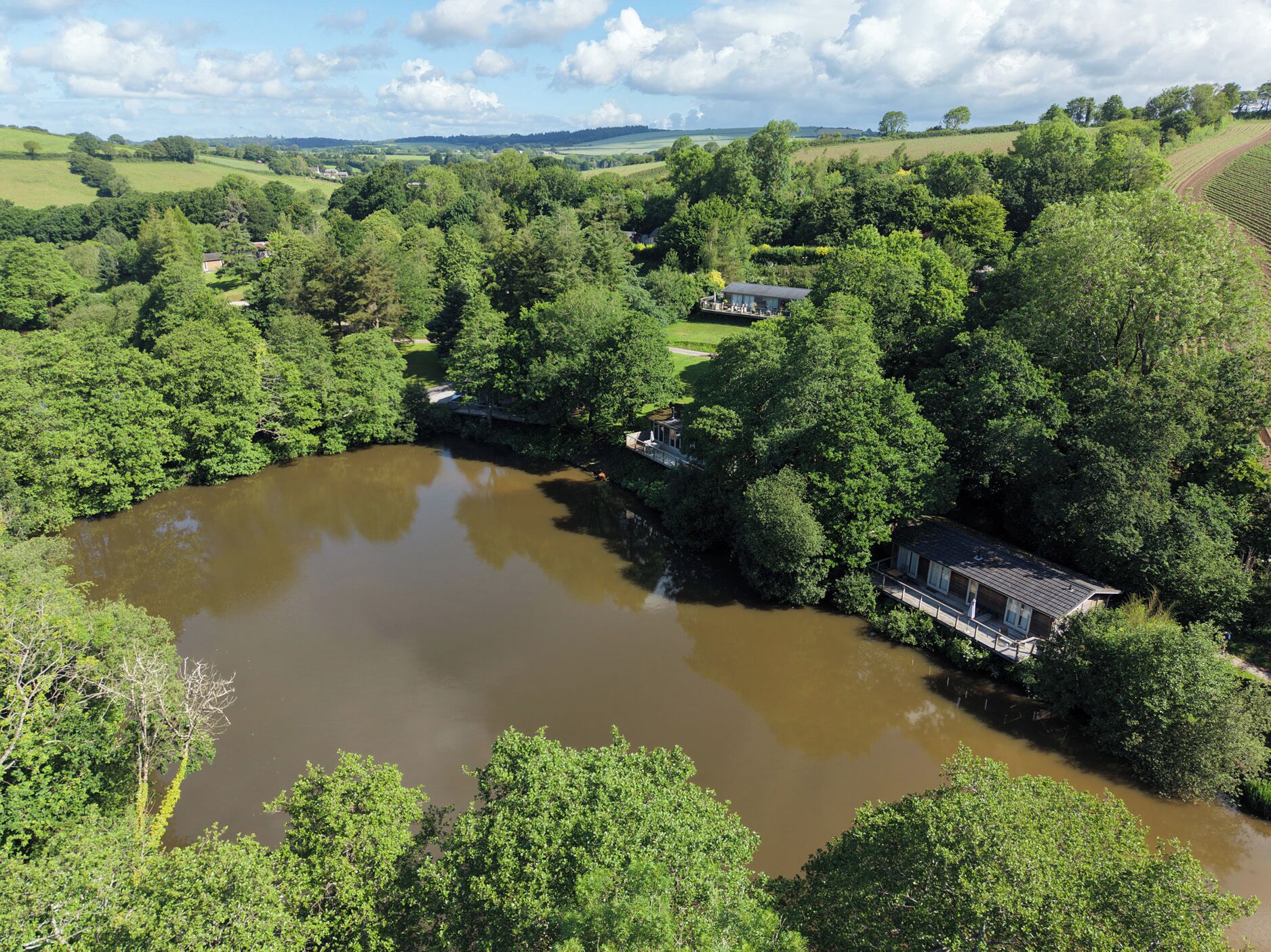 A riverside property with multiple buildings, including a house and a guesthouse, surrounded by lush greenery and a large body of water.