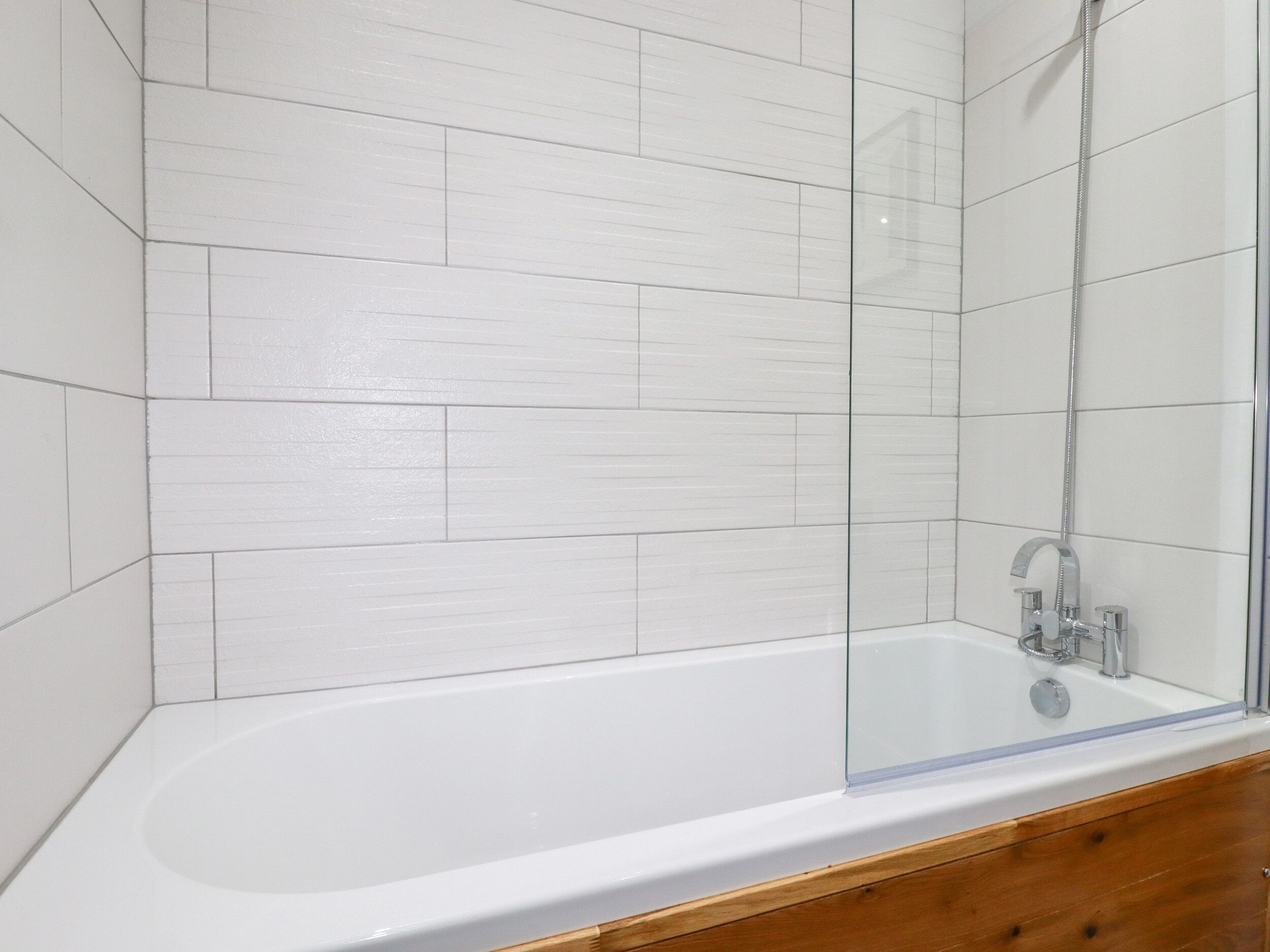 A modern bathroom with white tiled walls, a glass-enclosed bathtub, and a wooden vanity.