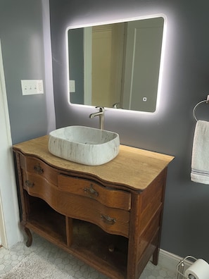 A bathroom with a marble sink, wooden vanity, a mirror with lights, a towel rack, and a toilet paper holder.